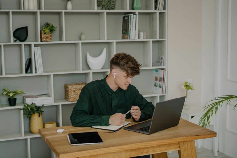 A Male Student in Green Long Sleeves Using a Laptop on a Wooden Desk A-Male-Student-in-Green-Long-Sleeves-Using-a-Laptop-on-a-Wooden-Desk-scaled