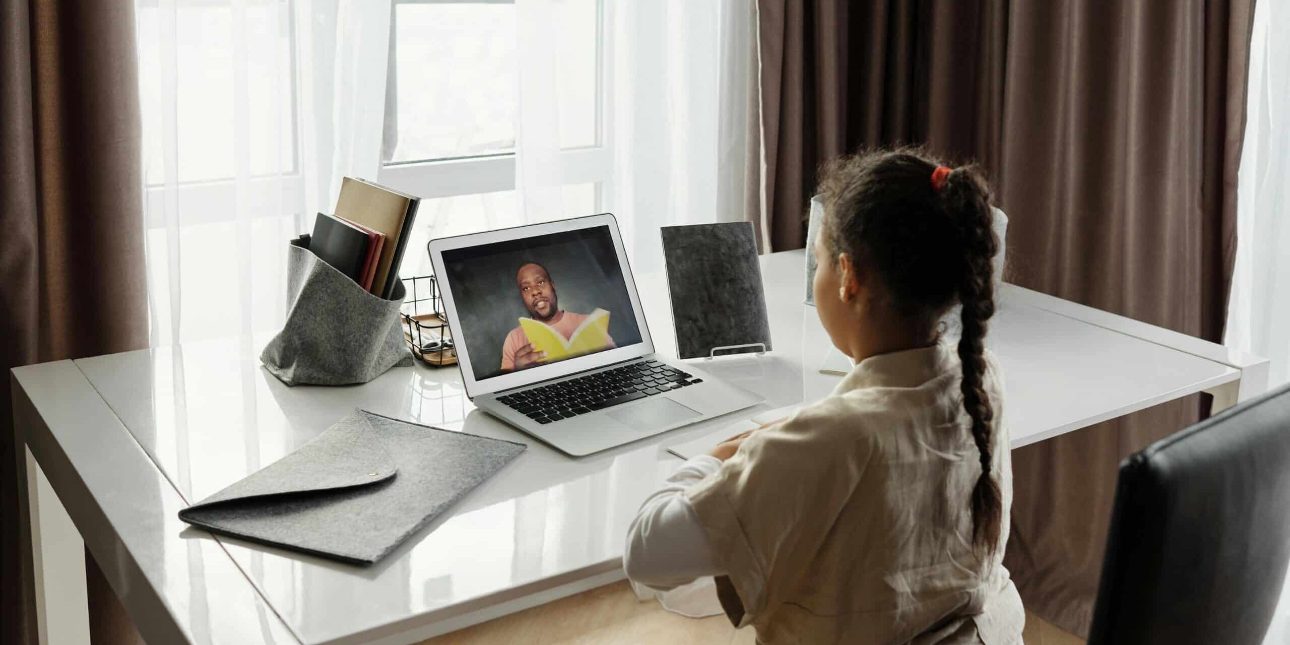 Child attending an online lesson on a laptop at a desk, watching a teacher on screen in a bright home setting.