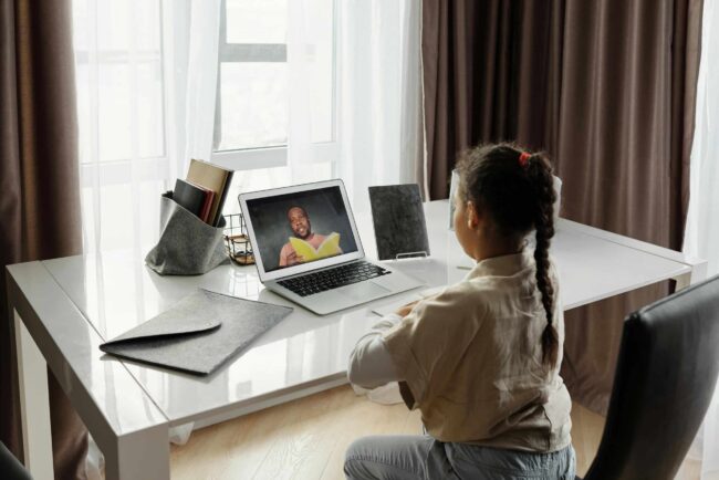 Child attending an online lesson on a laptop at a desk, watching a teacher on screen in a bright home setting.