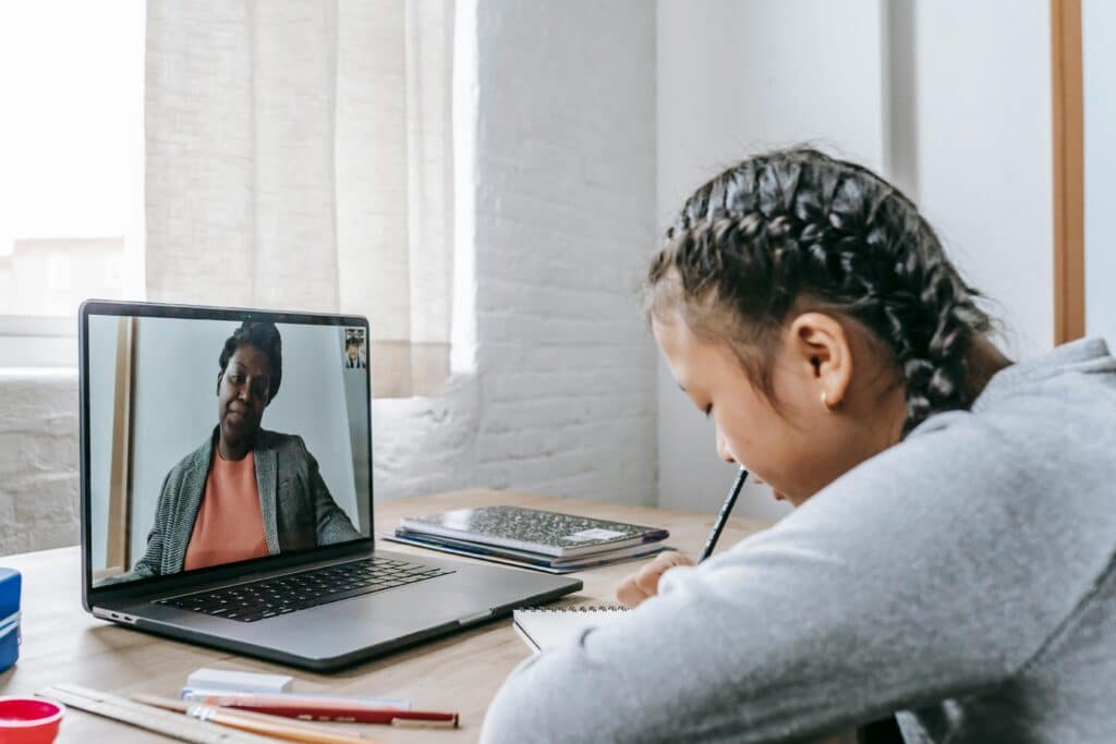 Student attending online class on laptop while writing notes at desk