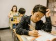 Student focused on writing in a workbook at a classroom desk, with classmates working quietly in the background.