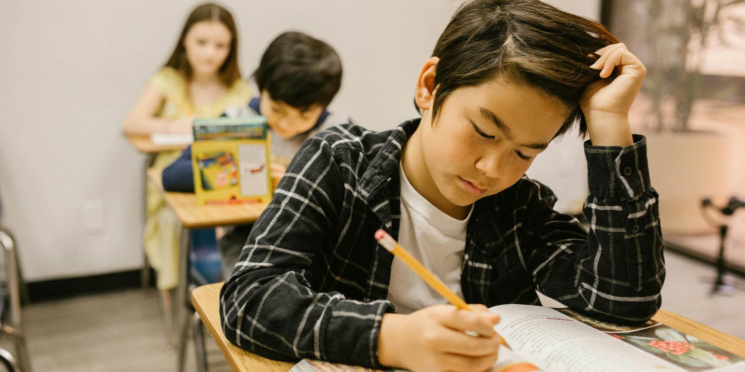 Student focused on writing in a workbook at a classroom desk, with classmates working quietly in the background.