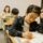 Student focused on writing in a workbook at a classroom desk, with classmates working quietly in the background.