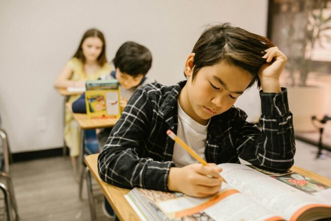 Student focused on writing in a workbook at a classroom desk, with classmates working quietly in the background.